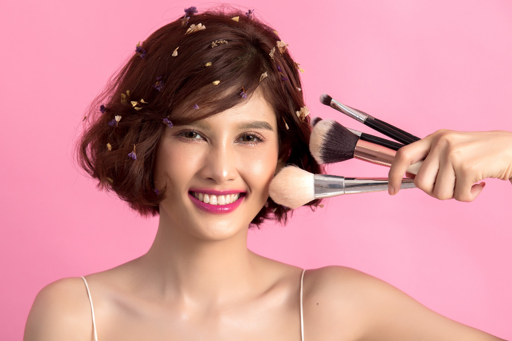 Modelo sonriente aplicando maquillaje con brocha grande sobre fondo rosa